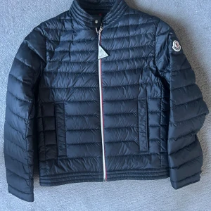 Svart dunjacka från Moncler - Snygg svart dunjacka från Moncler med quiltad design och dragkedja i kontrastfärg. Jackan har en hög krage och Moncler-logga på ärmen. Perfekt för kyliga dagar.