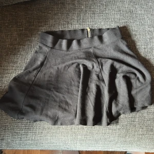 Svart kortkjol från Divided XS - Svart kortkjol från Divided i storlek XS. Kjolen har en snygg utsvängd form och en synlig dragkedja bak i guld. Tillverkad i mjuk bomullsblandning med polyester och elastan för extra stretch. Perfekt för dig som gillar en enkel men stilren look.