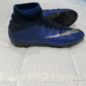 Blå Nike CR7 fotbollsskor med socka - Snygga blå fotbollsskor från Nike med CR7-detalj på hälen. Skorna har en hög stickad socka för extra stöd runt vristen och en silverfärgad swoosh på sidan. Ovandelen är i syntetmaterial och sulan har fasta dobbar för gräsplan. Fråga mig frågor och ge prisförslag 