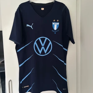 Malmö FF matchtröja  - Malmö FF fotbollströja i marinblått med ljusblå detaljer som är köpt i MFF shopen i Malmö, klubbmärke och två stjärnor på bröstet. Stort Wolkswagen-tryck på magen och Puma-logga på bröst och ärmar. Tillverkad i funktionsmaterial (Drycell) och har V-ringning samt korta ärmar. 