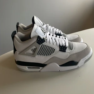 Nike Air Jordan 4 military black - Nike Air Jordan 4 sneakers i vitt och svart med klassisk meshpanel, Jumpman-logga på plösen och hälen samt coola detaljer i svart. Skorna har snörning, platt sula och är tillverkade i skinn och syntet. Perfekta för dig som vill sticka ut med en ikonisk siluett.