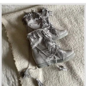 Grå mockaboots med fransar - Snygga grå boots i mocka med coola fransdetaljer och snörning. Skorna har platt sula och rund tå, perfekt för dig som vill sticka ut med en unik stil. Fransarna ger en boho-vibe och mockan känns lyxig och mjuk. Några så fläckar men inget som syns när man har dem på sig! 
