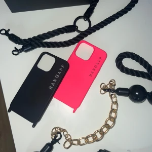 Designer-mobilskal från Hangapp (iPhone 13 PRO) - Trendiga och unika designer-mobildskal från märket Hangapp, den ena i färgen svart och den andra i neonrosa. Två grovt tvinnade svart rep att fästa i skalen, den ena lite lite längre med stora svarta pärldetaljer och kort guldkedja (avtagbar). Den andra mer simpel. Perfekt för att bära mobilen med stil och att mixa accessoarer. Originalpris på skalen är 300 kr st, och repen är värda 450 kr st. Guldkedjan värd 200 kr. Det rosa skalet är oanvänt och det svarta har ytterst små tecken på användning.