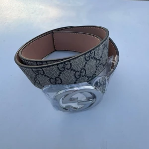Gucci GG Supreme bälte med silverdetalj - Helt oanvänd och snygg Gucci-bälte med klassiskt GG Supreme-mönster i beige och grått. Bältet har en bred design och en stor, rund spänne i silver med dubbla G-loggan. Tillverkat i skinn och canvas, perfekt för att lyfta vilken outfit som helst.