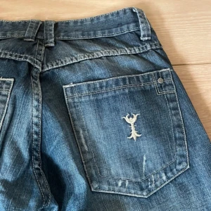  Affliction jeans blå - Säljer ett par i blå denim med coola patchade detaljer på benen och broderad logga på bakfickan. Klassisk femficksmodell med raka ben och normal passform. Perfekta för dig som gillar en unik streetstil. W32 L34