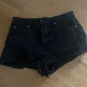 Svarta jeansshorts med fransig kant - Snygga svarta jeansshorts med råa, fransiga kanter och slitna detaljer. Klassisk femficksmodell med knapp och dragkedja framtill. Perfekta för en avslappnad och cool stil under varma dagar.