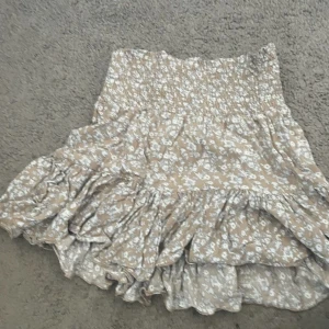 Beige blommig volangkjol - Säljer en supersöt kort kjol i beige med vitt blommönster och volanger. Säljer då den inte kommer till användning✨💕.