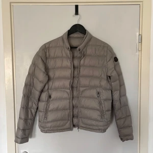 Beige Moncler dunjacka med dragkedja - Snygg beige dunjacka från Moncler med klassisk quiltad design och hög krage. Jackan har två fickor med dragkedja och Moncler-logga på ärmen. Perfekt för kyliga dagar och ger en clean, stilren look.