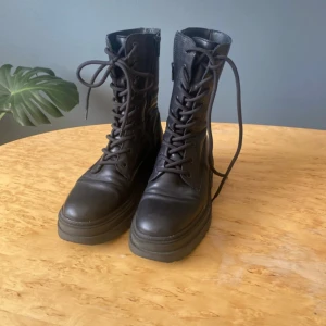 Svarta chunky boots med snörning - Säljer ett par svarta boots i skinn med grov platt sula och snörning framtill. Skorna har dragkedja på sidan för enkel påtagning och en rund tå. Perfekta för dig som gillar en edgy och stilren look. Originalpris , ca 1000kr.