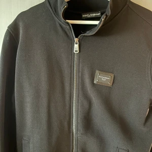 tröja från DOLCE & GABBANA - Svart kofta från Dolce & Gabbana med klassisk siluett och snygg metallogo på bröstet. Jackan har dragkedja framtill, ståkrage och diskreta fickor. Materialet är kraftigt och ger en exklusiv känsla. Perfekt för dig som gillar stilrena och lyxiga plagg.