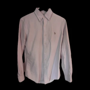 Ljusrosa skjorta från Polo Ralph Lauren - Säljer en klassisk ljusrosa skjorta från Polo Ralph Lauren. Skjortan har lång ärm, knappar hela vägen och den ikoniska broderade loggan på bröstet. Tillverkad i mjuk bomull och har en snygg, tidlös passform med krage.