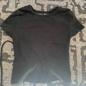 Svart topp med snörning från Cropp - Cool svart topp från Cropp med korta ärmar och unik snörningsdetalj i ryggen som ger en edgy vibe. Gjord i mjuk bomull och perfekt för dig som vill sticka ut lite extra. Enkel framtill och öppen rygg med snygg knytning.