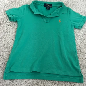 Grön pikétröja från Ralph Lauren - Snygg grön pikétröja från Polo Ralph Lauren med klassisk krage och korta ärmar. Tröjan har den ikoniska orange broderade loggan på bröstet och är tillverkad i mjuk bomull. Perfekt för en chill och stilren look. För barn