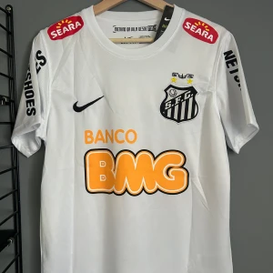Santos Neymar Jr 11 Nike fotbollströja - Vit Santos fotbollströja från Nike med Neymar Jr och nummer 11 på ryggen. Tröjan har korta ärmar, klubbmärke på bröstet och flera sponsorloggor i svart, orange och rött. Tillverkad i lätt och ventilerande material, perfekt för match eller träning. Helt ny med tagg en replika 