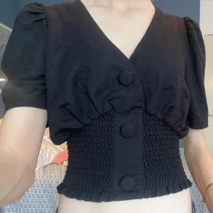 Svart blus med smock från Gina Tricot - Supersnygg svart blus från Gina Tricot med puffärmar, v-ringning och stora klädda knappar framtill. Midjan har smockdetalj som ger en tight och croppad passform! 💕Köpt på Plick och bara använd 1 gång :)
