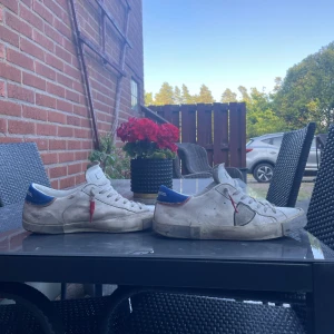 Philippe Model vita sneakers med blå detalj - Snygga vita sneakers från Philippe Model Paris med blå hälkappa och röda detaljer på sidan. Klassisk låg modell med snörning och rund tå. Skorna har en platt sula och är tillverkade i skinn för en exklusiv känsla.