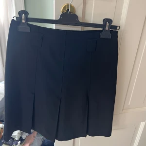 Svart kortkjol från Sinéquanone - Stilren svart kortkjol från Sinéquanone med snygga veck framtill. Kjolen har en klassisk rak passform och breda hällor i midjan. Perfekt att matcha med en cool topp eller oversized jacka.