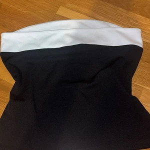 Svart och vit bandeau tubtopp - Snygg bandeau tubtopp i svart med bred vit kant upptill. Toppen är axelbandslös och har en tight passform som sitter snyggt mot kroppen. Perfekt att styla med höga byxor eller kjol för en clean och modern look.