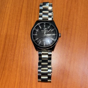 Emporio Armani silverfärgad klocka - Stilren klocka från Emporio Armani med svart urtavla och markerade index. Armbandet är i blankt silverfärgat metall och boetten har en klassisk rund form. Urtavlan har dag- och datumvisning samt diskret mönster. Perfekt accessoar för en snygg och lyxig look. Skriv vid minsta fundering. Pris är ej hugget i sten🙌🏼