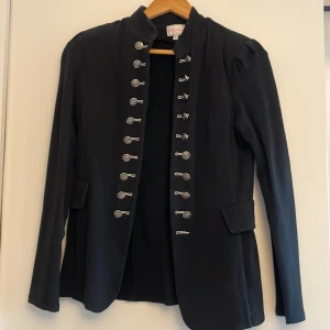 Svart rock från Chloe Underhill - Snygg svart rock från Chica London  med coola silvriga knappar i två rader framtill och ståkrage. Jackan har långa ärmar, fickdetaljer och en rak passform. Perfekt för dig som vill sticka ut med en militärinspirerad look.