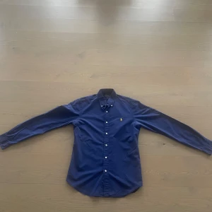Marinblå skjorta Polo Ralph Lauren M - Snygg marinblå skjorta från Polo Ralph Lauren i slim fit-modell. Klassisk design med knappar hela vägen, lång ärm och ikonisk gul broderad logga på bröstet. Skjortan är tillverkad i bomull och passar perfekt till en stilren look. Ny pris 1800kr