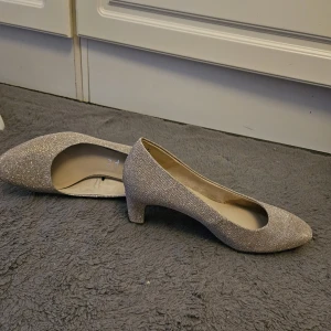 Tamaris Pumps Guld Storlek 39 Toppskick - Eleganta guldiga pumps från Tamaris i storlek 39. Perfekta för festliga tillfällen eller för att piffa upp vardagsoutfiten. Bekväm klackhöjd som gör dem lätta att bära hela kvällen. Enda deffekten är sulan under. Annars finns det inget slitagepåverkan varken på klacken eller skon. Använd 2 ggr