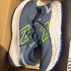 Blå New Balance sneakers med Fresh Foam - Snygga blå sneakers från New Balance med Fresh Foam-sula för extra komfort. Ovandelen är i mesh med grå och neongula detaljer, perfekt för dig som gillar sportig stil. Skorna har snörning och en tjock, vit sula med blå undersida.