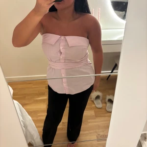 Rosa randig bandeau tubtopp med knappar - Säljer en unik bandeau tubtopp i ljusrosa och vit med tunna ränder. Toppen har knappar framtill, en kragdetalj upptill och ett brett band som markerar midjan. Perfekt för dig som vill sticka ut med en kreativ och trendig look.