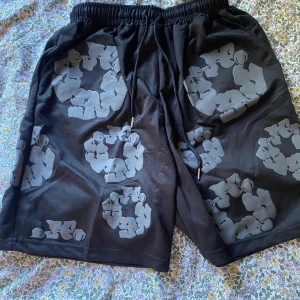 Svarta shorts från Denim Tears - Svarta shorts från Denim Tears med unikt grått blommönster över hela plagget. De har elastisk midja med snörning och en bakficka. Perfekta för en chill och trendig sommarstil.