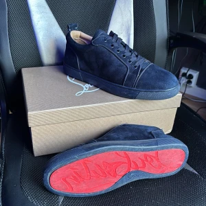 Mörkblå mockasneakers från Louboutin - Exklusiva mörkblå sneakers i mocka från Christian Louboutin med ikonisk röd sula och broderad logga på hälen. Skorna har snörning och rund tå, samt en platt sula som ger en clean och lyxig vibe. Perfekta för dig som vill sticka ut med stil.