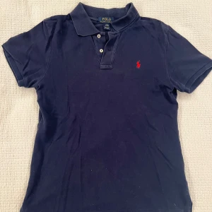 Marinblå pikétröja från Ralph Lauren - Klassisk marinblå pikétröja från Ralph Lauren med korta ärmar och krage. Tröjan har två knappar framtill och den ikoniska röda logotypen broderad på bröstet. Tillverkad i mjuk bomull som är skön att bära hela dagen.