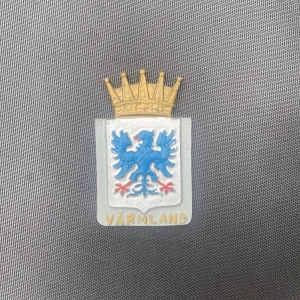 Värmland vapensköld brosch - Cool brosch i form av Värmlands vapensköld med blå grip, guldkrona och texten 'Värmland' i guld. Skölden är i silverfärg med detaljer i blått, rött och guld. Perfekt accessoar för dig som vill visa ditt ursprung eller gillar heraldik.