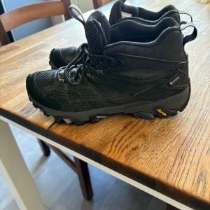 Svarta Merrell kängor med Gore-Tex - Svarta kängor från Merrell med Gore-Tex-membran och robust Vibram-sula. Skorna har snörning, meshpaneler för andning och förstärkta partier i mocka och syntet. Perfekta för utomhusaktiviteter och äventyr.