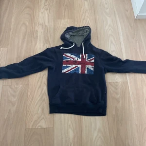 Mörkblå hoodie med Union Jack-tryck - Säljer en mörkblå hoodie med Union Jack-flagga på bröstet. Tröjan har huva med vita snören och en stor magficka. Perfekt för dig som gillar streetstyle och vill sticka ut lite extra. Skön passform och långärmad modell.