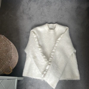 Cos tröja stickad me volang armar  - Superfin vit stickad tröja från cos  med volangdetaljer diagonalt över framsidan. Tröjan har  långa ärmar, perfekt för att hålla sig varm och snygg under kyliga dagar. Mjuk och fluffig känsla som ger en mysig vibe.