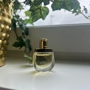 Elegant parfymflaska med gulddetaljer - Snygg parfymflaska i genomskinligt glas med rundad form och lyxiga gulddetaljer på korken. Perfekt att ha framme på sminkbordet för en exklusiv känsla. Innehållet är ljust och flaskan känns modern och stilren.