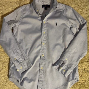 Ljusblå skjorta från Ralph Lauren - Klassisk ljusblå skjorta från Ralph Lauren med broderad logga på bröstet. Skjortan har lång ärm, knappar hela vägen och button down-krage. Snygg passform och tillverkad i bomull, perfekt för en clean och stilren look.