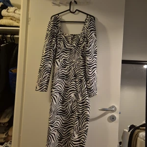 Zebramönstrad långklänning Gina Tricot - Snygg långklänning från Gina Tricot med zebramönster i svart och vitt. Klänningen har långa ärmar, fyrkantig ringning och en slits på sidan för extra edge. Perfekt för dig som vill sticka ut med ett djurmönster och cool siluett.