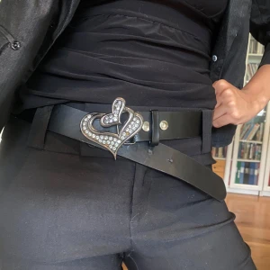 Svart bälte med glittrigt hjärtspänne - Säljer ett svart bälte i skinn med ett stort, glittrigt hjärtformat spänne dekorerat med stenar. Bältet har en klassisk form och passar perfekt för att ge outfiten en cool och unik touch. Perfekt accessoar för dig som gillar statement-detaljer.