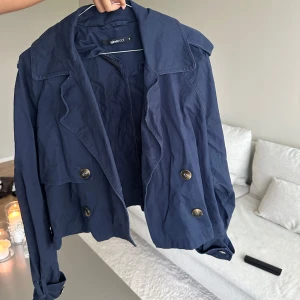 Mörkblå kort trenchcoat från Ginatricot - Kort trenchcoat i mörkblått från Ginatricot med stora slag, dubbelknäppning och snygga axeldetaljer. Jackan har långa ärmar och är tillverkad i bomull, 💙