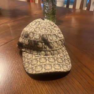Beige keps från Salvatore Ferragamo - Snygg keps från Salvatore Ferragamo i beige och svart med ikoniskt mönster över hela kepsen. Justerbart spänne bak och böjd skärm. Tillverkad i ett slitstarkt tyg med exklusiv känsla, perfekt för dig som vill sticka ut med designeraccessoar.