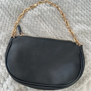 Svart axelväska från MNG med guldkedja - Snygg svart axelväska från MNG i skinnimitation med guldfärgad kedja som axelrem. Väskan har rundad form och dragkedja upptill. Perfekt storlek för en clean look till din outfit.