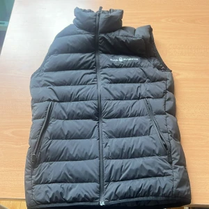 Svart dunväst från Sail Racing - Snygg svart dunväst från Sail Racing med quiltad design och hög krage. Västen har dragkedja framtill och två praktiska fickor med dragkedja. Perfekt för lager-på-lager och stilren look. Logga på bröstet och nacken för extra detalj.