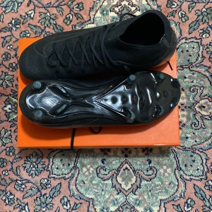 Svarta Nike fotbollsskor med snörning - Säljer ett par helsvarta Nike fotbollsskor med hög krage och snörning. Skorna har en mönstrad ovandel i syntetmaterial och fasta dobbar för gräsplan. Snygg, slimmad design som ger bra grepp och kontroll på planen.
