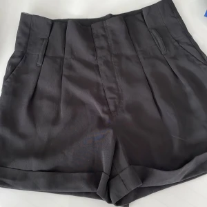 Svarta högmidjade shorts från Bik Bok - Stilrena svarta shorts från Bik Bok i högmidjad modell med uppvikta benslut och pressveck framtill. Shortsen har hällor för bälte och diskreta fickor bak. Perfekta för en clean och snygg look under varmare dagar.