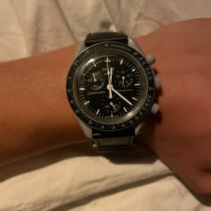 Omega x Swatch MoonSwatch Speedmaster - Säljer en Omega x Swatch MoonSwatch Speedmaster med svart urtavla, vita visare och detaljer samt svart textilband. Klockan har en rund boett i biokeramik och klassisk tachymeter-ring. Perfekt för dig som gillar ikonisk design och rymdinspirerade accessoarer. 