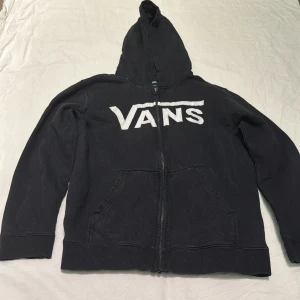 Svart hoodie från Vans med logga - Svart hoodie från Vans i storlek L med stor vit Vans-logga framtill. Tröjan har dragkedja, huva och två fickor på magen. Perfekt för en avslappnad streetstil och funkar året runt. Tillverkad i mjuk bomullsmix för skön känsla.