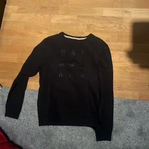 Svart sweatshirt från Blend, storlek L - Svart sweatshirt från Blend i storlek L. Tröjan har rund halsringning, långa ärmar och tryck med svarta bokstäver framtill. Tillverkad i mjuk bomullsmix som är skön att ha på sig. Perfekt för en chill och stilren look.