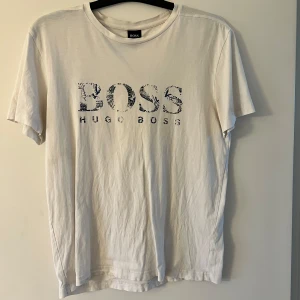Vit t-shirt från Hugo Boss - Vit t-shirt från Hugo Boss med svart tryck på bröstet där det står BOSS HUGO BOSS. Klassisk passform med rund hals och korta ärmar. Perfekt för en clean och stilren look.