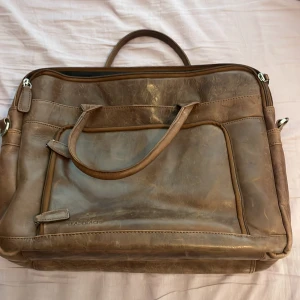 Brun skinn portfölj  - Snygg portfölj i brunt skinn från Saddler med flera fack och dragkedjor. Klassisk design med dubbla handtag och detaljer i silverfärgad metall. Perfekt för laptop och dokument, med rymligt innerfack och smarta fickor på utsidan.